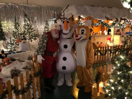 Santa Claus poses with a snowman and a reindeer mascot in a festive, decorated holiday setting.