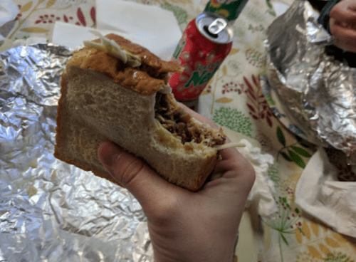 A hand holding a partially eaten sandwich with a can of soda in the background, on a patterned tablecloth.