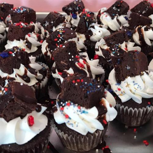 A close-up of chocolate cupcakes topped with whipped cream, chocolate drizzle, and sprinkles, featuring brownie pieces.