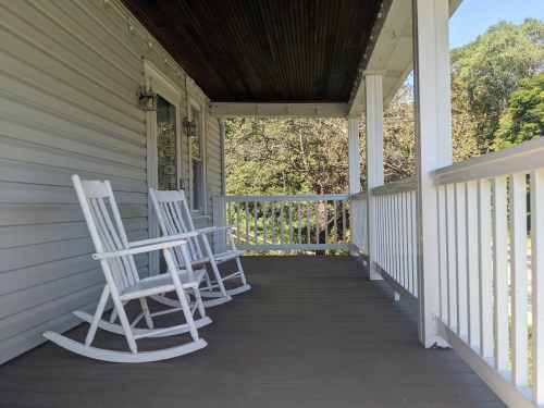 Two white rocking chairs on a porch with a wooden floor and a view of greenery in the background.