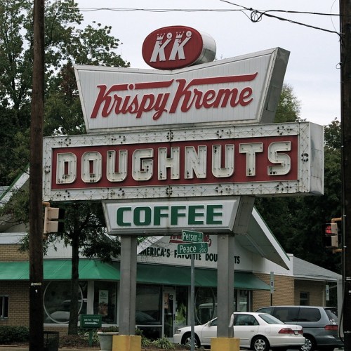 Krispy Kreme doughnut shop sign featuring "Doughnuts" and "Coffee" in bold letters, surrounded by trees and buildings.
