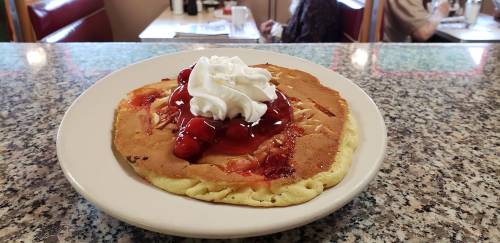 A plate of pancakes topped with whipped cream and cherry sauce, set on a granite countertop in a diner.