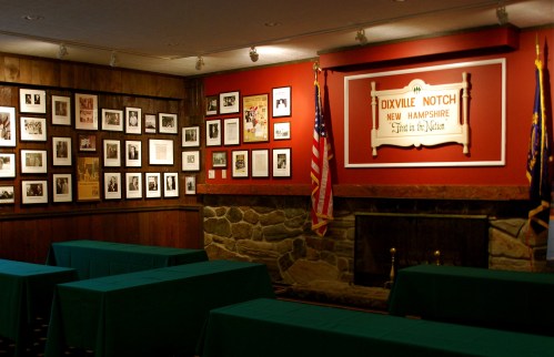 Interior of a room with green tables, a stone fireplace, and walls adorned with historical photos and a sign for Dixville Notch.