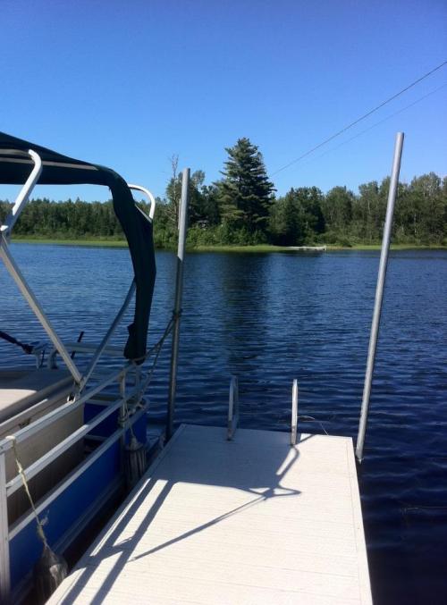 A peaceful lakeside scene featuring a dock, a boat, and lush green trees under a clear blue sky.