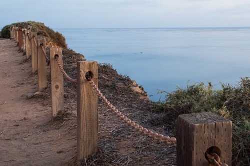 A wooden fence with a chain along a coastal path, overlooking calm waters and a cloudy sky.