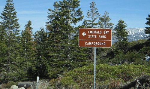 Brown sign pointing left to Emerald Bay State Park Campground, surrounded by tall green trees and mountains in the background.