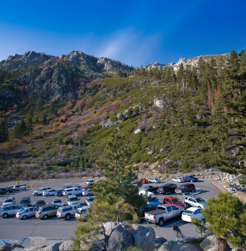 A parking lot filled with cars, surrounded by mountains and trees under a clear blue sky.