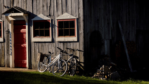 A rustic barn with a red door, featuring two white bicycles parked outside and a pile of wood nearby.