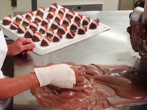 A hand in a glove spreads melted chocolate on a surface, with chocolate-covered strawberries in the background.