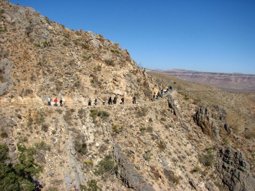A winding trail on a rocky hillside with hikers walking along the path under a clear blue sky.