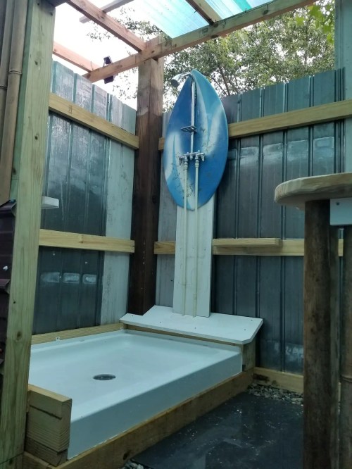 Outdoor shower area with wooden walls, a surfboard mounted above, and a white shower base on a stone floor.