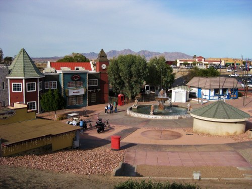 A scenic view of a small town square featuring a fountain, shops, and mountains in the background under a clear sky.