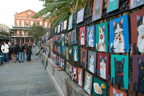 A vibrant street scene featuring a wall of colorful cat paintings, with people browsing nearby in an outdoor setting.
