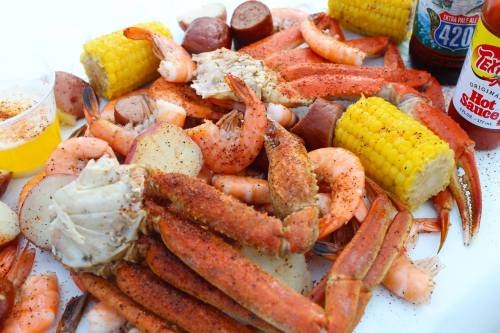 A colorful seafood platter featuring shrimp, crab, sausage, corn, and potatoes, with hot sauce and a drink on the side.