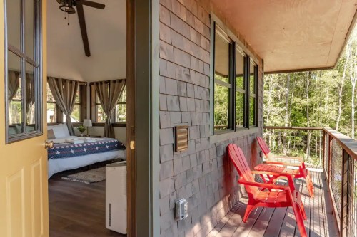 A cozy porch with two red chairs overlooks a forested area, leading to a well-decorated interior space.