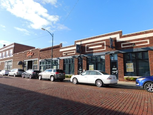 A row of brick buildings with storefronts, parked cars along a cobblestone street, under a clear blue sky.