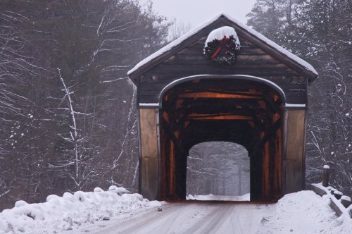 A snow-covered road leads through a wooden covered bridge adorned with a Christmas wreath.