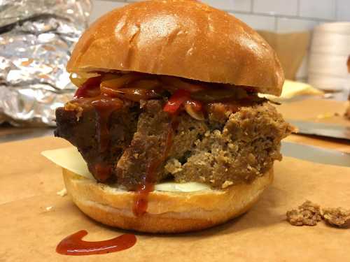 A hearty meatloaf sandwich on a bun, topped with ketchup, onions, and cheese, served on a brown paper surface.