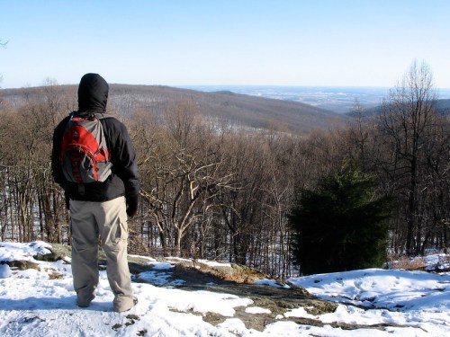 A hiker in winter gear stands on a rocky ledge, overlooking a snowy landscape and distant hills.