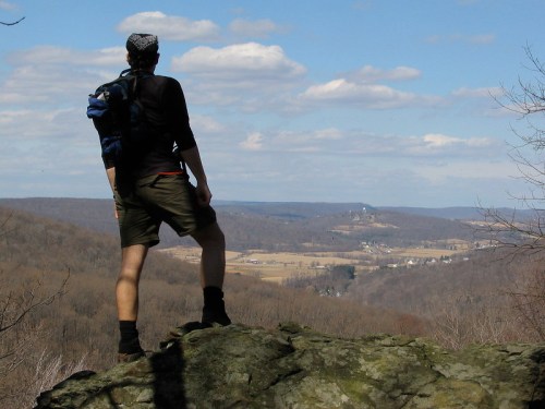 A hiker stands on a rock, gazing at a valley and distant hills under a partly cloudy sky.