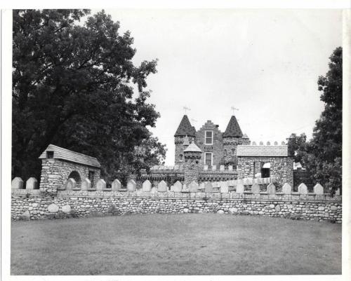 A stone castle with turrets surrounded by a low wall and trees, captured in black and white.