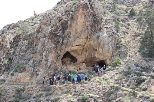 A group of people gathers near a rocky cave entrance on a hillside, surrounded by sparse vegetation.