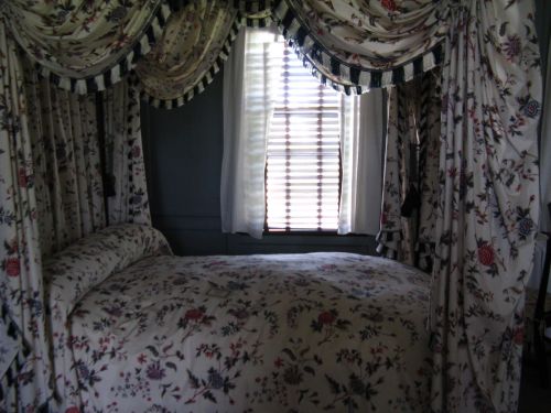 A cozy bedroom with a floral canopy bed, draped curtains, and a window with blinds letting in soft light.