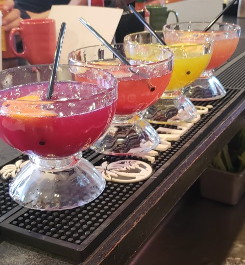 Four colorful cocktails in clear bowls, garnished with fruit, lined up on a bar counter.