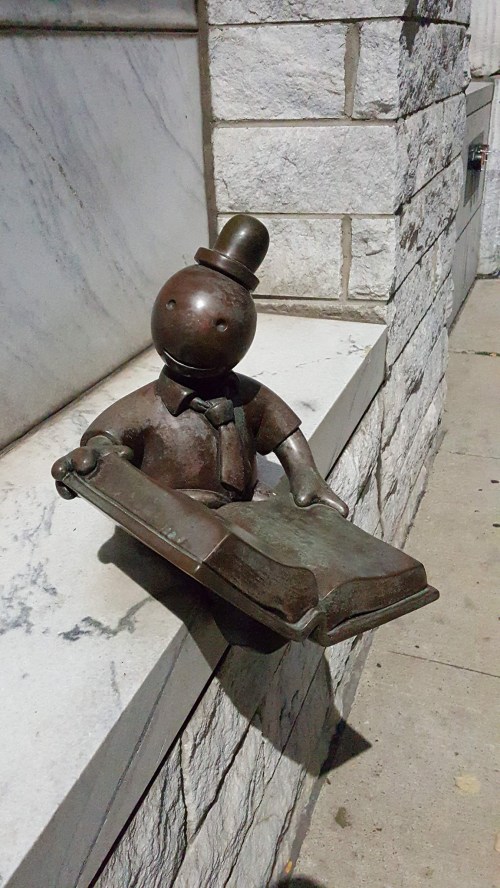 A bronze statue of a character in a hat, sitting on a ledge, holding a book.