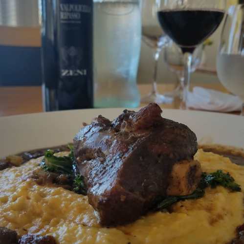 A plate of tender meat on creamy polenta, garnished with greens, with wine glasses and a bottle in the background.