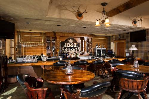 A rustic saloon interior featuring a wooden bar, round tables, and a variety of drinks displayed on shelves.
