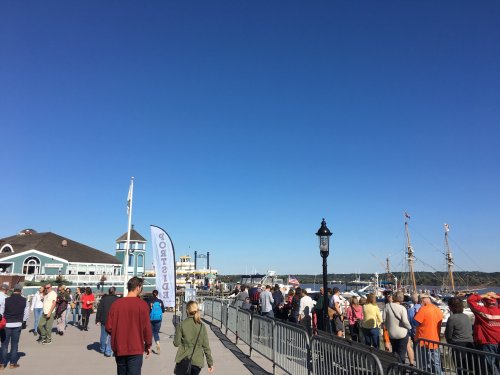 A busy waterfront scene with people walking along a boardwalk, boats in the harbor, and clear blue skies.