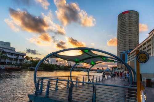 A waterfront promenade with a curved canopy, overlooking a river at sunset, with clouds and a tall building in the background.