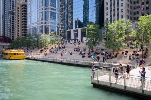 A vibrant riverside scene with people relaxing on steps, a yellow boat, and tall buildings reflecting in the water.