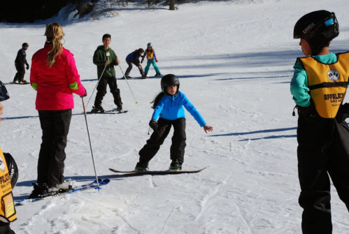 A group of children skiing on a snowy slope, with one child practicing their skills while others watch.