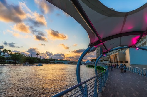 A scenic waterfront walkway with a colorful sunset, featuring people strolling along the riverbank.
