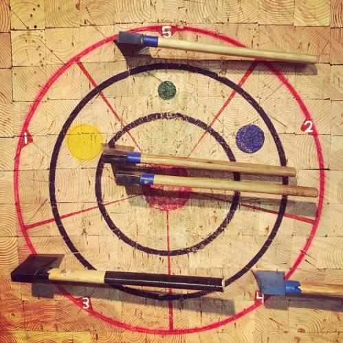A wooden target board with colored circles and numbered sections, featuring several axes stuck in various positions.
