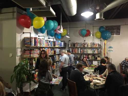 A lively game night in a cozy space with colorful balloons, shelves of board games, and people enjoying activities.