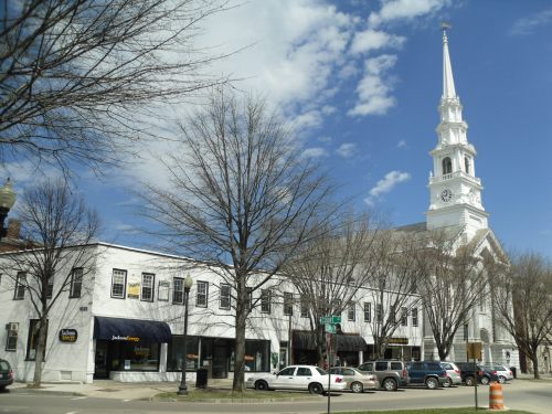A white church with a tall steeple beside a row of shops, under a blue sky with scattered clouds.
