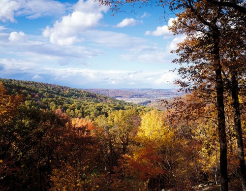 A scenic view of rolling hills covered in vibrant autumn foliage under a partly cloudy sky.