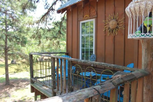 A wooden balcony with blue chairs overlooks a green yard, featuring a sun decoration on the wall and trees in the background.