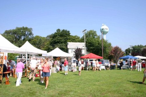 A bustling outdoor market with vendors under tents, people browsing, and a water tower in the background on a sunny day.