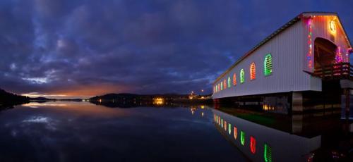 A covered bridge adorned with colorful lights reflects in calm water at dusk, surrounded by a scenic landscape.
