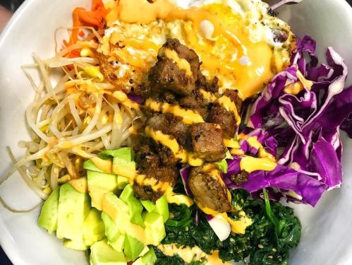A colorful bowl of rice topped with vegetables, avocado, fried egg, and seasoned meat drizzled with sauce.