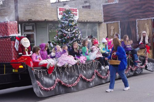 A festive parade float with children in colorful outfits, a Christmas tree, and Santa decorations, passing by a woman.