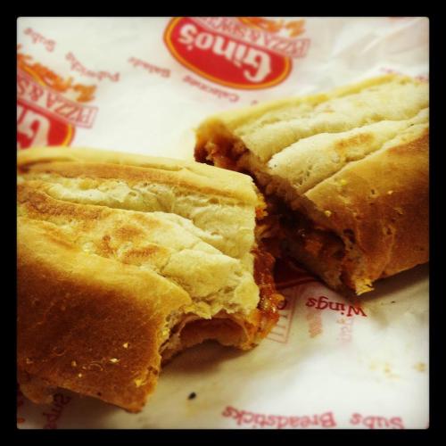A close-up of a partially eaten sandwich with a soft bread roll and filling, placed on a branded paper wrapper.