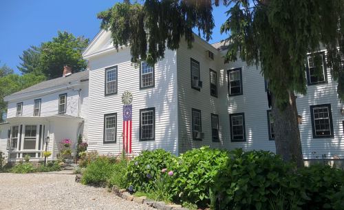 A white house with black shutters, an American flag, and a garden, set against a clear blue sky.