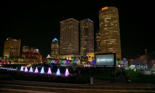 City skyline at night with colorful lights, a large screen, and people gathered in a park.