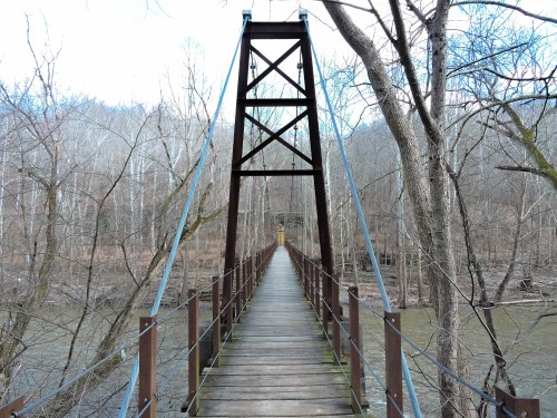 A suspension bridge spans a river, surrounded by bare trees and a serene landscape in early spring.