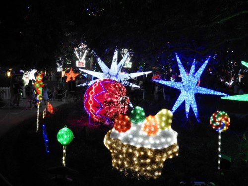Colorful holiday lights in various shapes, including stars, ornaments, and a dessert, illuminate a festive outdoor scene.